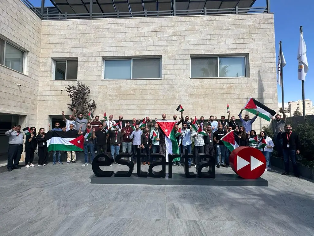 A group of people standing in front of the Estarta building, holding Jordanian flags and waving in solidarity. The Estarta logo is visible beside them. The building has a modern design with multiple windows.