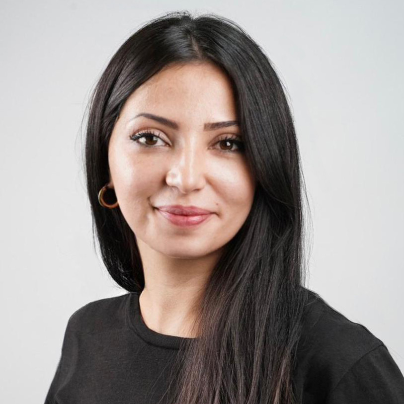 A woman with long, straight dark hair smiles slightly at the camera, perhaps considering her next step in her career. She is wearing a black shirt and gold hoop earrings. The background is plain and light-colored.