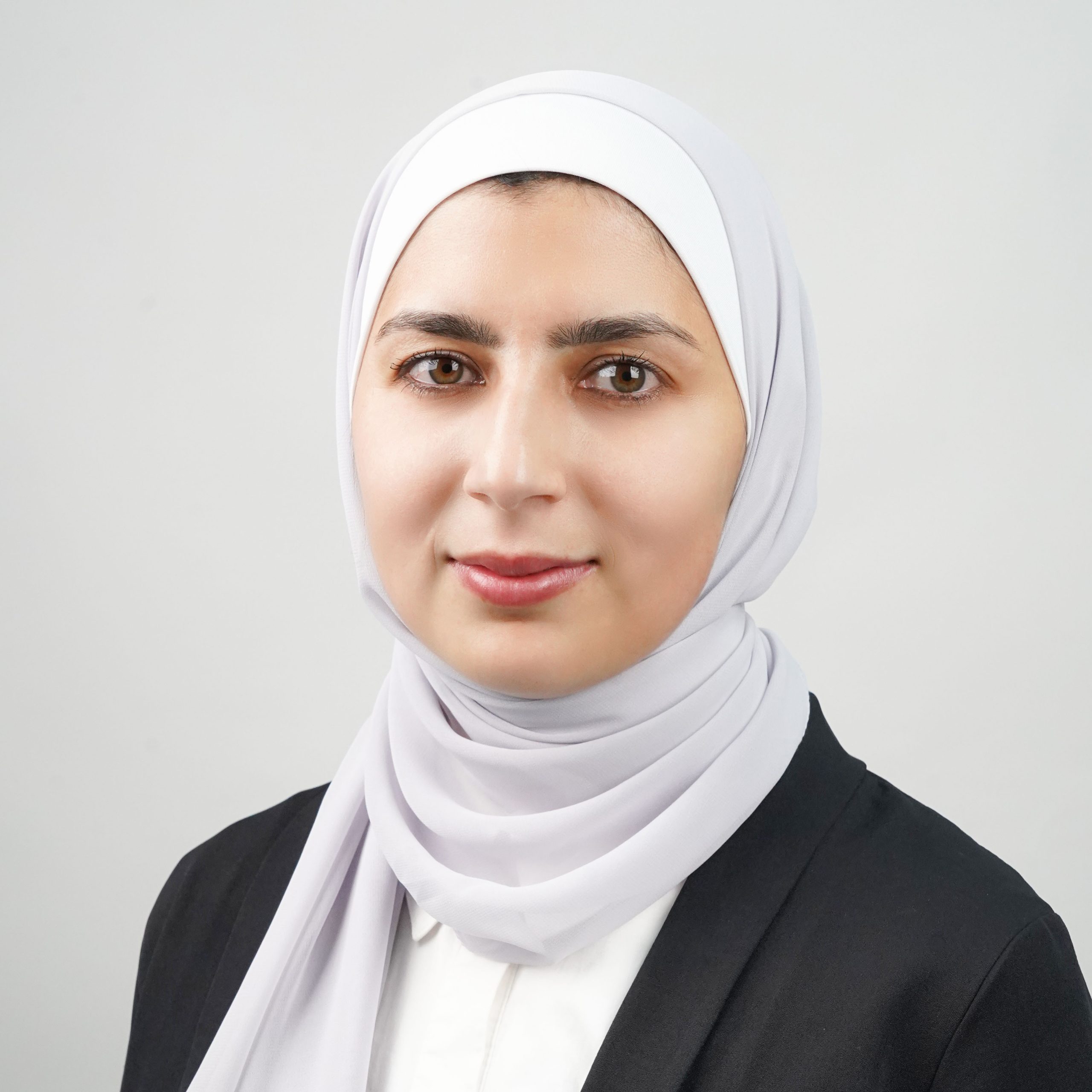 A woman wearing a light gray hijab, a white shirt, and a black blazer is looking directly at the camera. She has a neutral expression and is posed in front of a plain, light gray background, exuding professionalism perfect for careers in any field.