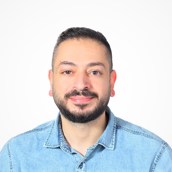 A man with short dark hair and a beard is smiling at the camera, exuding confidence in his light blue denim shirt. Set against a plain white background, he appears ready for new career opportunities.