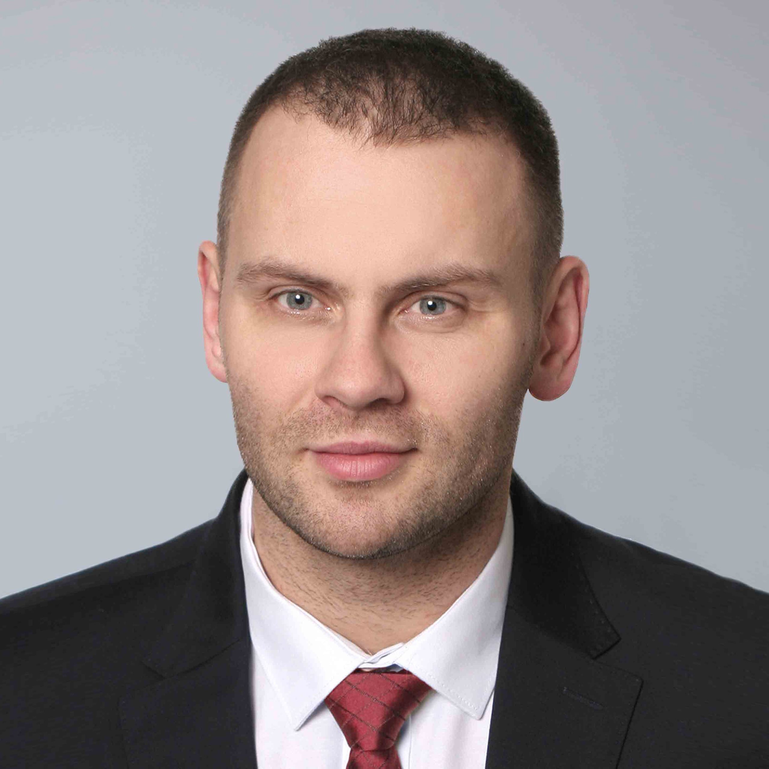 A professional portrait of a man with short brown hair, wearing a black suit, white shirt, and red tie. He has a slight stubble and is looking forward with a neutral expression. The light gray background emphasizes his poised demeanor, suitable for diverse careers.